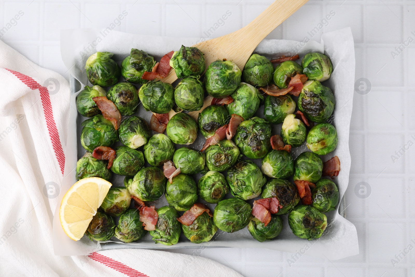 Photo of Delicious roasted Brussels sprouts, bacon, lemon and wooden spatula on white tiled table, top view
