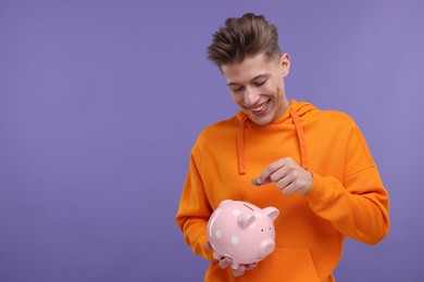Photo of Happy man putting coins into piggy bank on purple background. Space for text