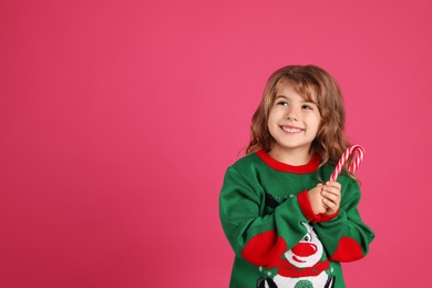 Photo of Cute little girl in Christmas sweater holding sweet candy cane against pink background. Space for text