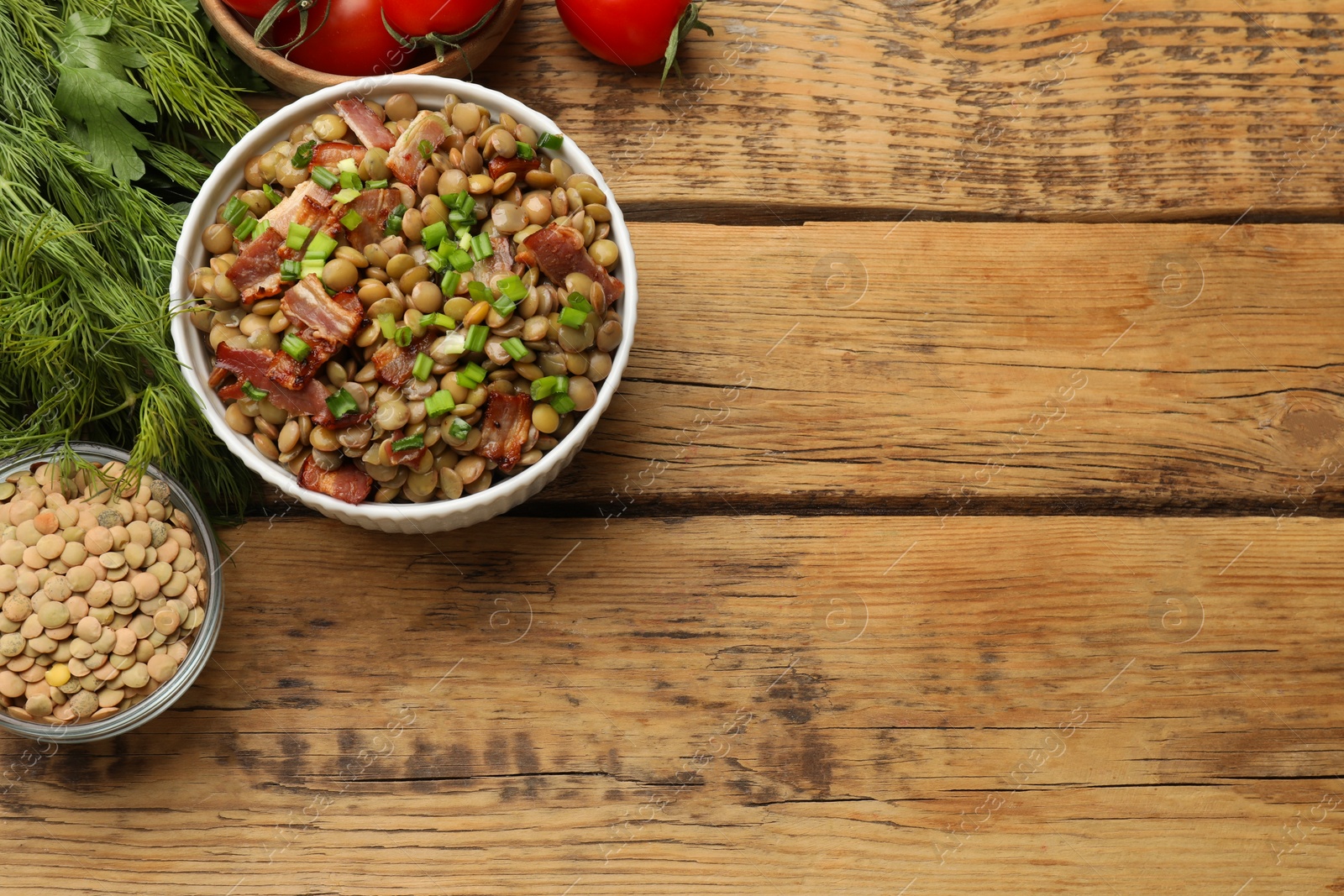 Photo of Delicious lentils with bacon, green onion in bowl, tomatoes and greens on wooden table, flat lay. Space for text