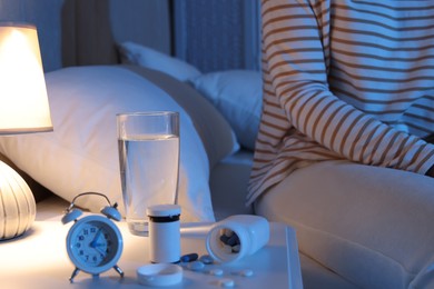 Woman near nightstand with pills, water and alarm clock in bedroom at night, closeup