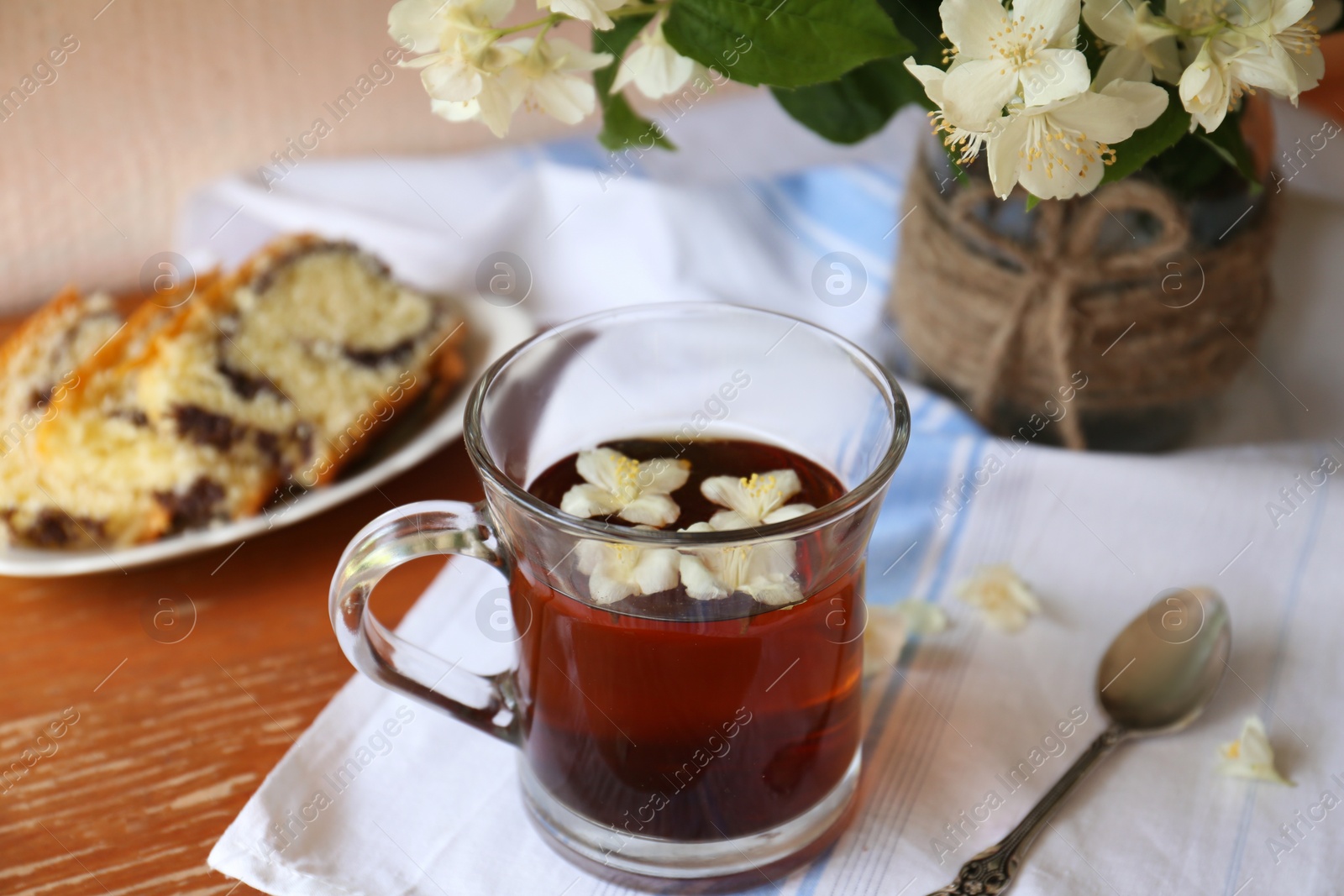 Photo of Glass cup of aromatic tea, tasty dessert and beautiful jasmine flowers on wooden table