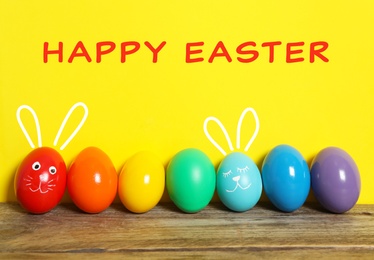 Two eggs with drawn faces and ears as Easter bunnies among others on wooden table against yellow background