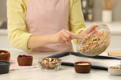 Photo of Making granola. Woman putting mixture of oat flakes, dried fruits and other ingredients onto baking tray at light table, closeup