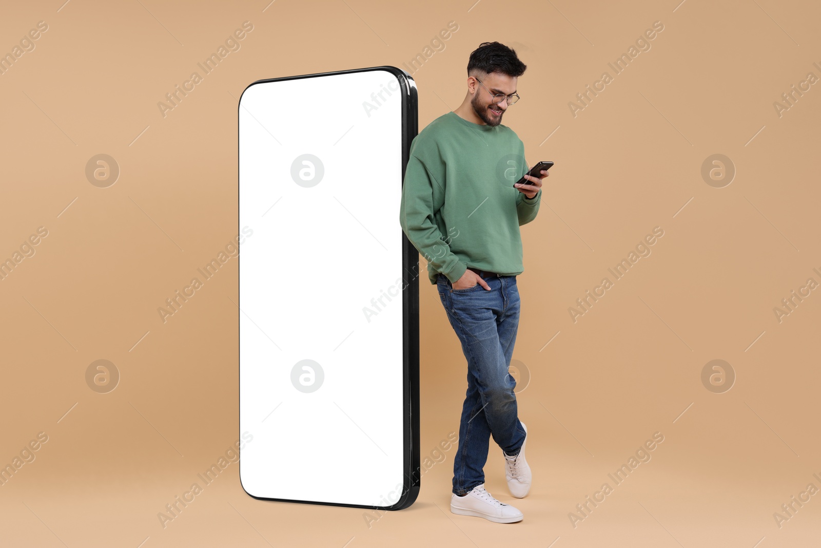 Image of Man with mobile phone standing near huge device with empty screen on dark beige background. Mockup for design