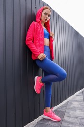 Beautiful woman in gym clothes posing near dark grey fence on street, low angle view