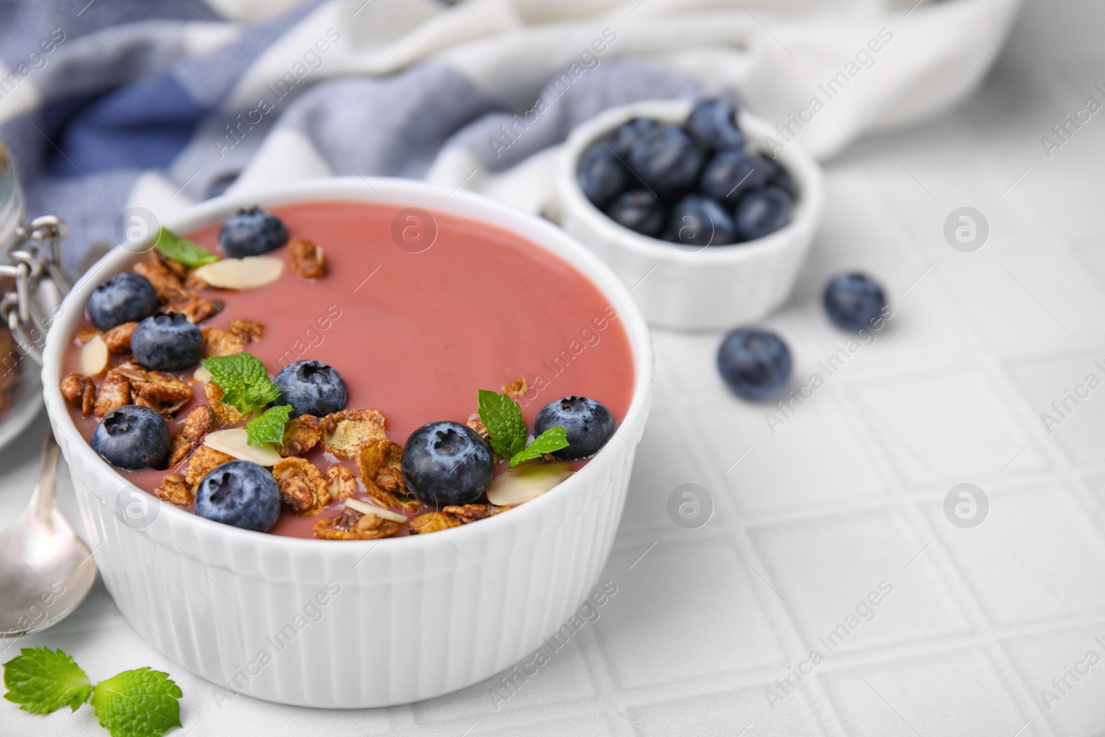 Photo of Bowl of delicious smoothie with fresh blueberries and granola on white tiled table, closeup. Space for text