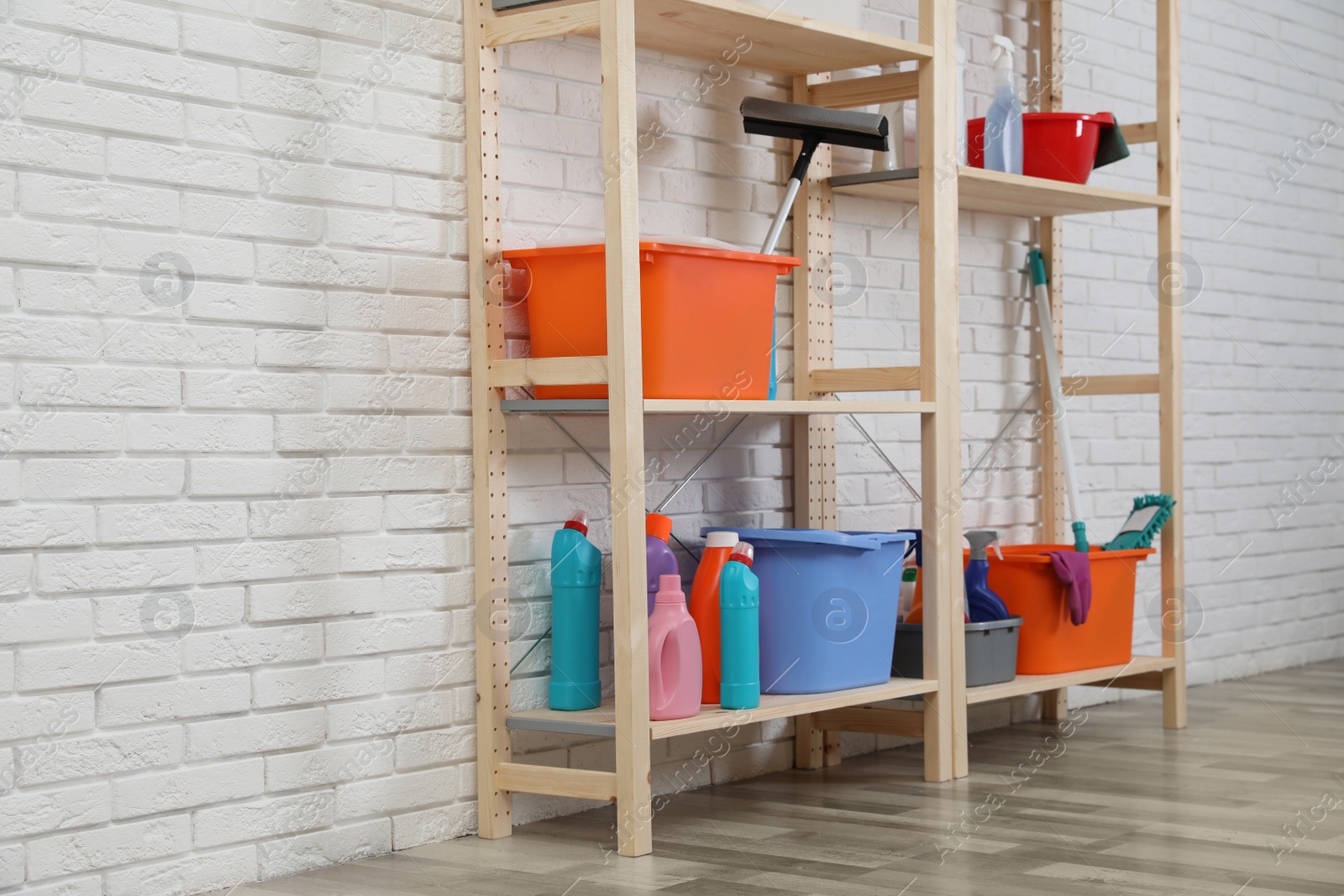 Photo of Wooden shelving units with cleaning equipment near white brick wall. Stylish room interior