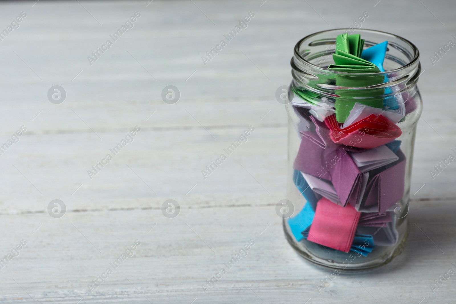 Photo of Glass jar with colorful paper pieces on white wooden table, space for text