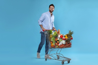 Photo of Young man with shopping cart full of groceries on light blue background