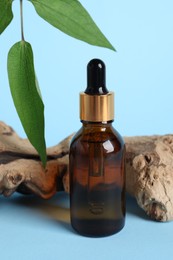 Bottle with cosmetic oil, green leaves and wooden snag on light blue background, closeup