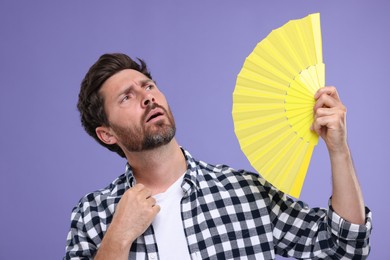 Unhappy man with hand fan suffering from heat on purple background