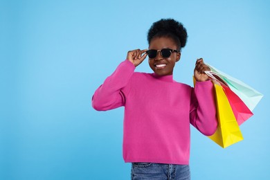 Happy young woman in stylish sunglasses with shopping bags on light blue background