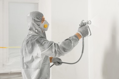 Decorator in protective overalls painting wall with spray gun indoors