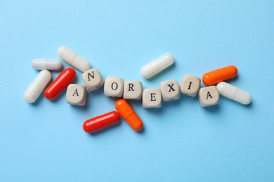 Photo of Word Anorexia of wooden cubes and pills on light blue background, flat lay