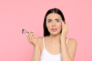 Emotional young woman with eyelash curler on light pink background