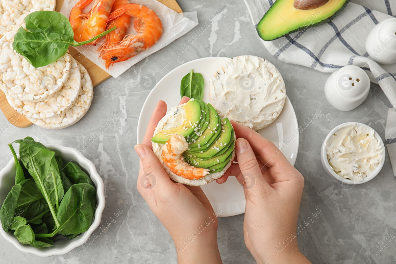 Photo of Woman holding puffed rice cake with shrimp and avocado at grey marble table, top view