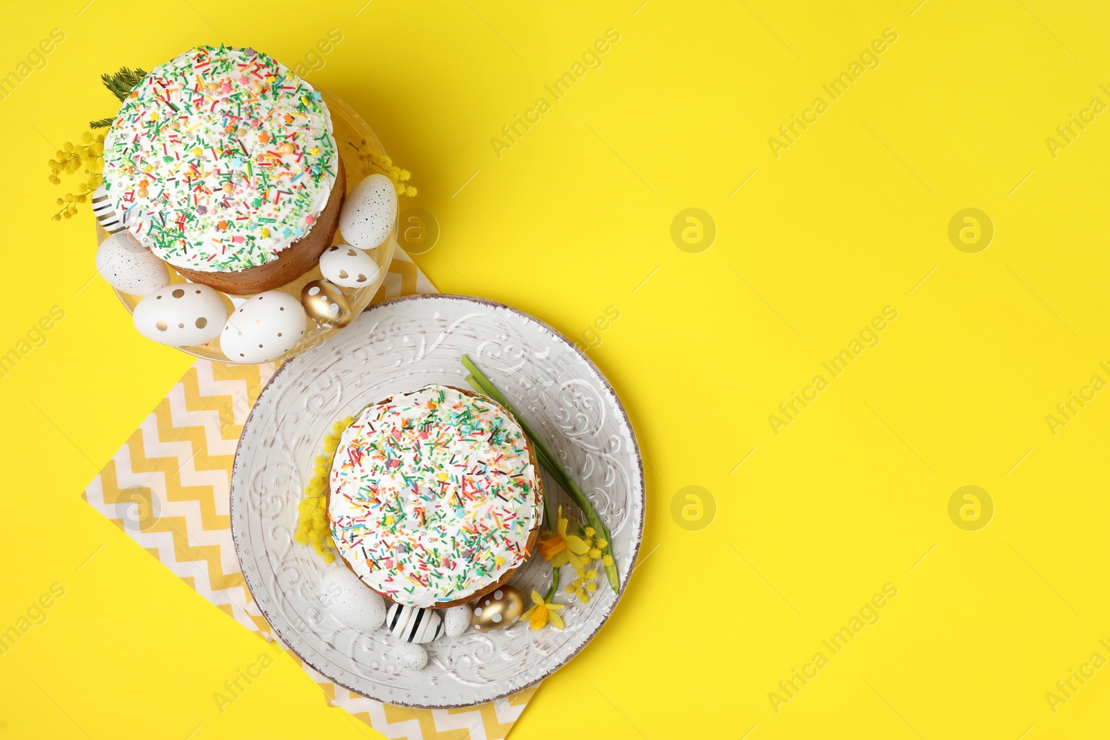 Photo of Traditional Easter cakes with sprinkles, painted eggs and beautiful spring flowers on yellow background, flat lay. Space for text