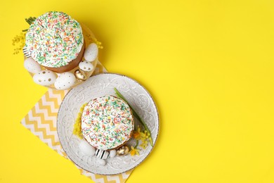 Photo of Traditional Easter cakes with sprinkles, painted eggs and beautiful spring flowers on yellow background, flat lay. Space for text