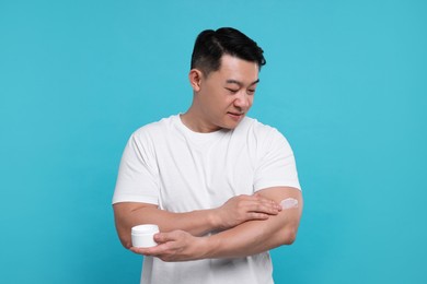 Photo of Handsome man applying body cream onto his arm on light blue background