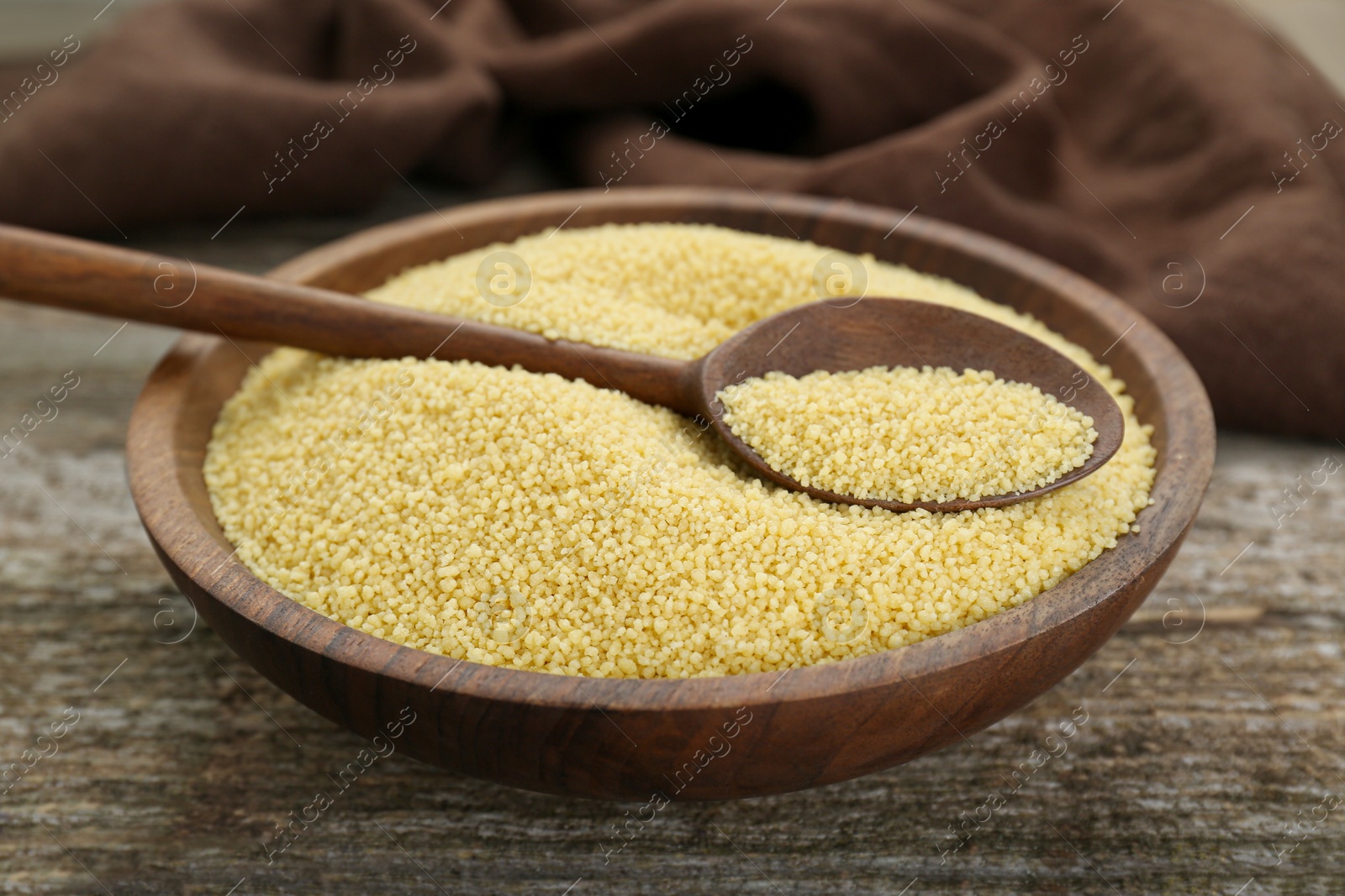 Photo of Bowl and spoon with raw couscous on wooden table, closeup