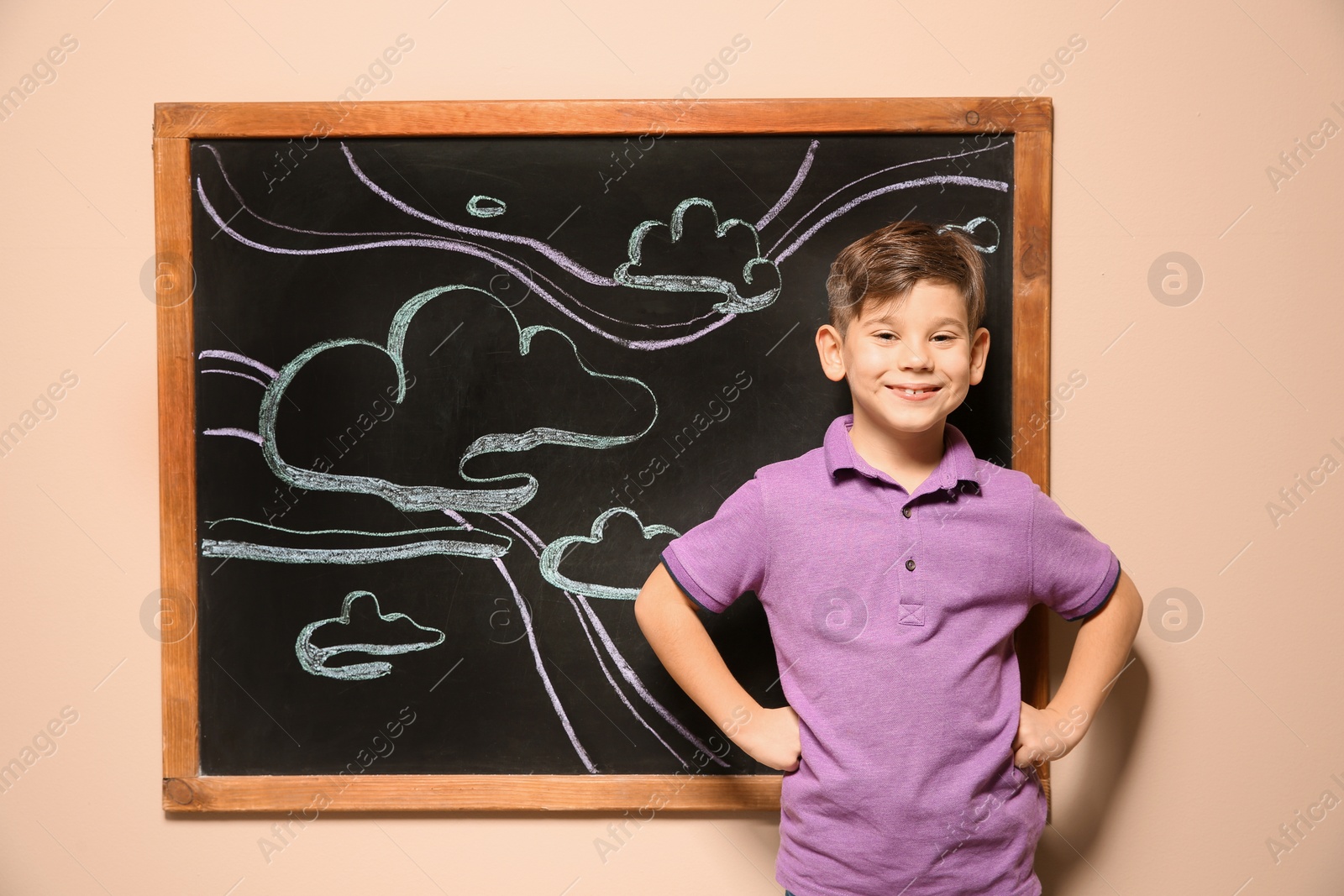 Photo of Cute little child standing at blackboard with chalk drawn sky and clouds