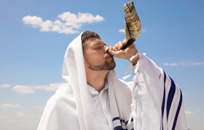 Photo of Jewish man in tallit blowing shofar outdoors. Rosh Hashanah celebration