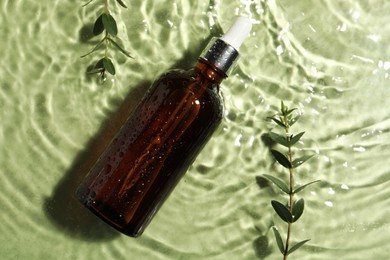 Bottle of hydrophilic oil and plant twigs in water on light green background, flat lay