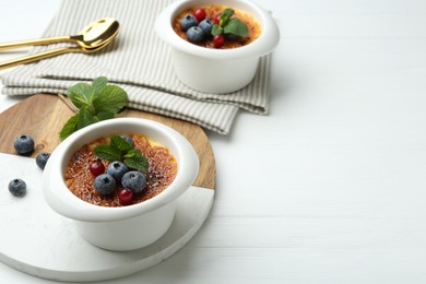 Photo of Delicious creme brulee with berries and mint in bowls on white wooden table, closeup. Space for text