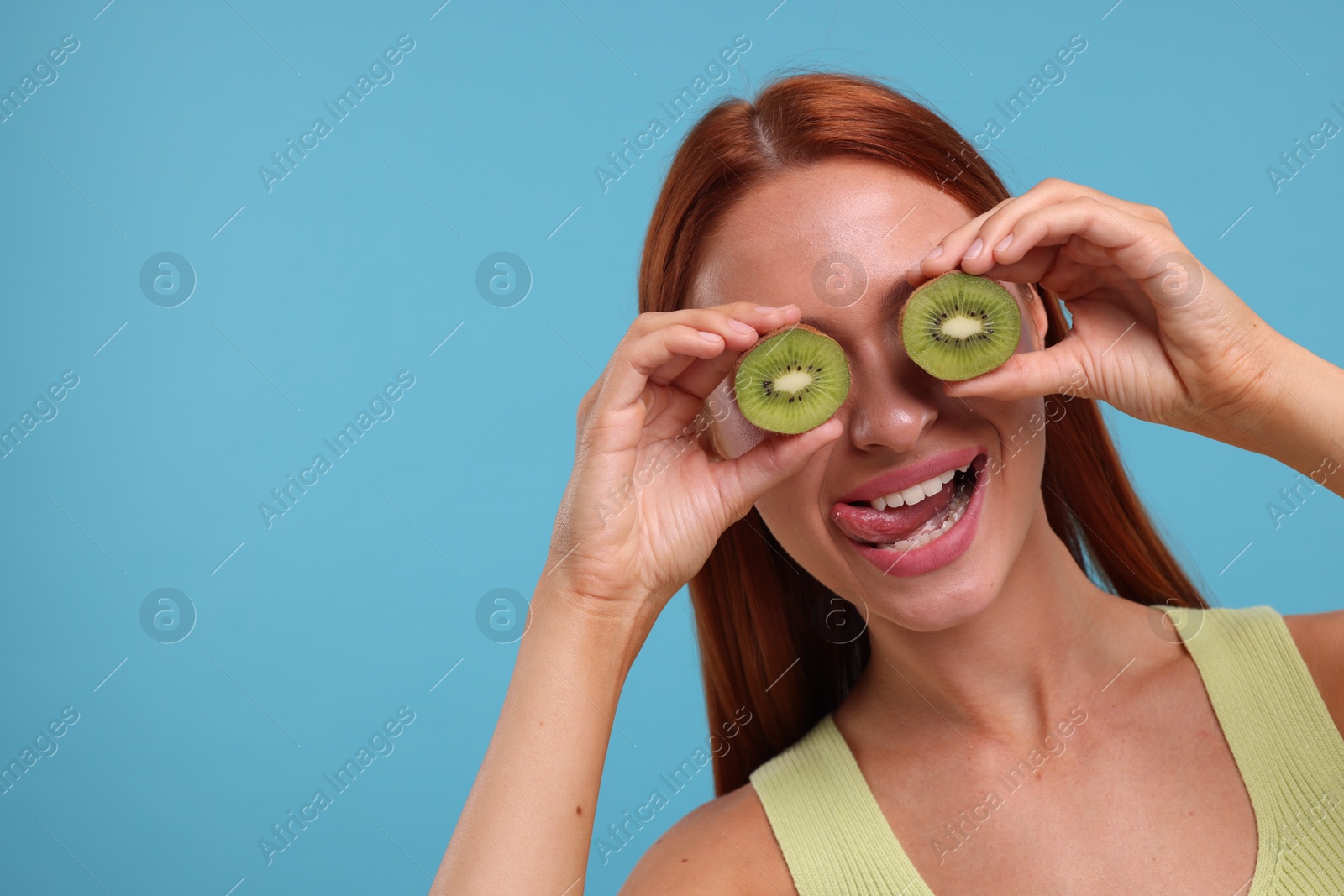 Photo of Funny woman covering eyes with halves of fresh kiwi and showing tongue on light blue background, space for text