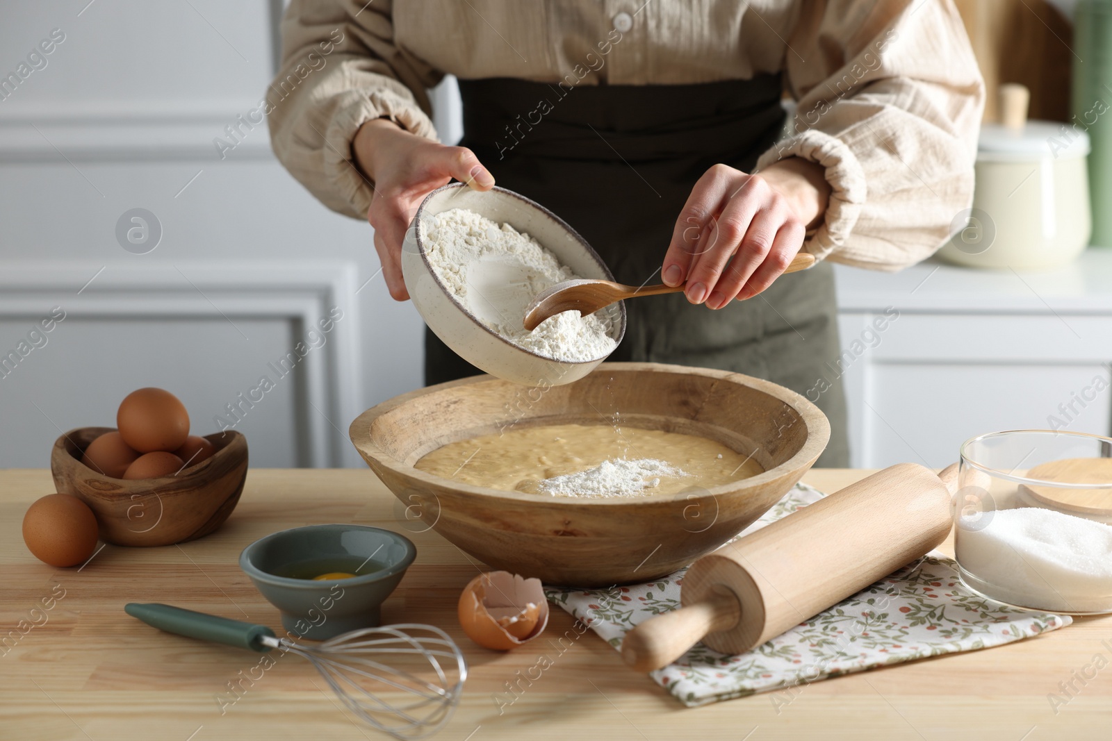 Photo of Making dough. Woman adding flour into bowl at wooden table in kitchen, closeup