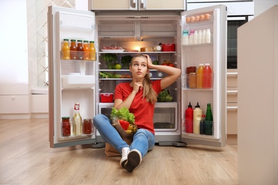 Photo of Young woman with paper bag full of food sitting near refrigerator in kitchen