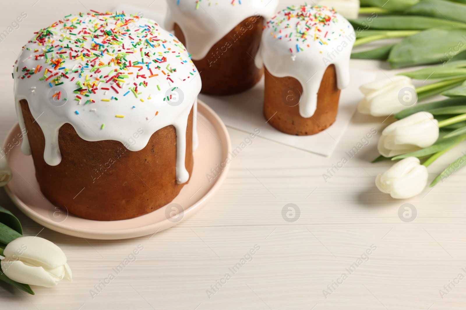 Photo of Delicious Easter cakes with sprinkles and beautiful tulips on white wooden table. Space for text