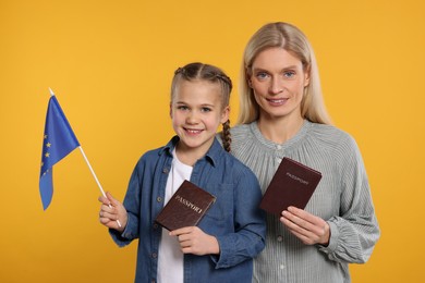 Photo of Immigration. Happy woman with her daughter holding passports and flag of European Union on orange background