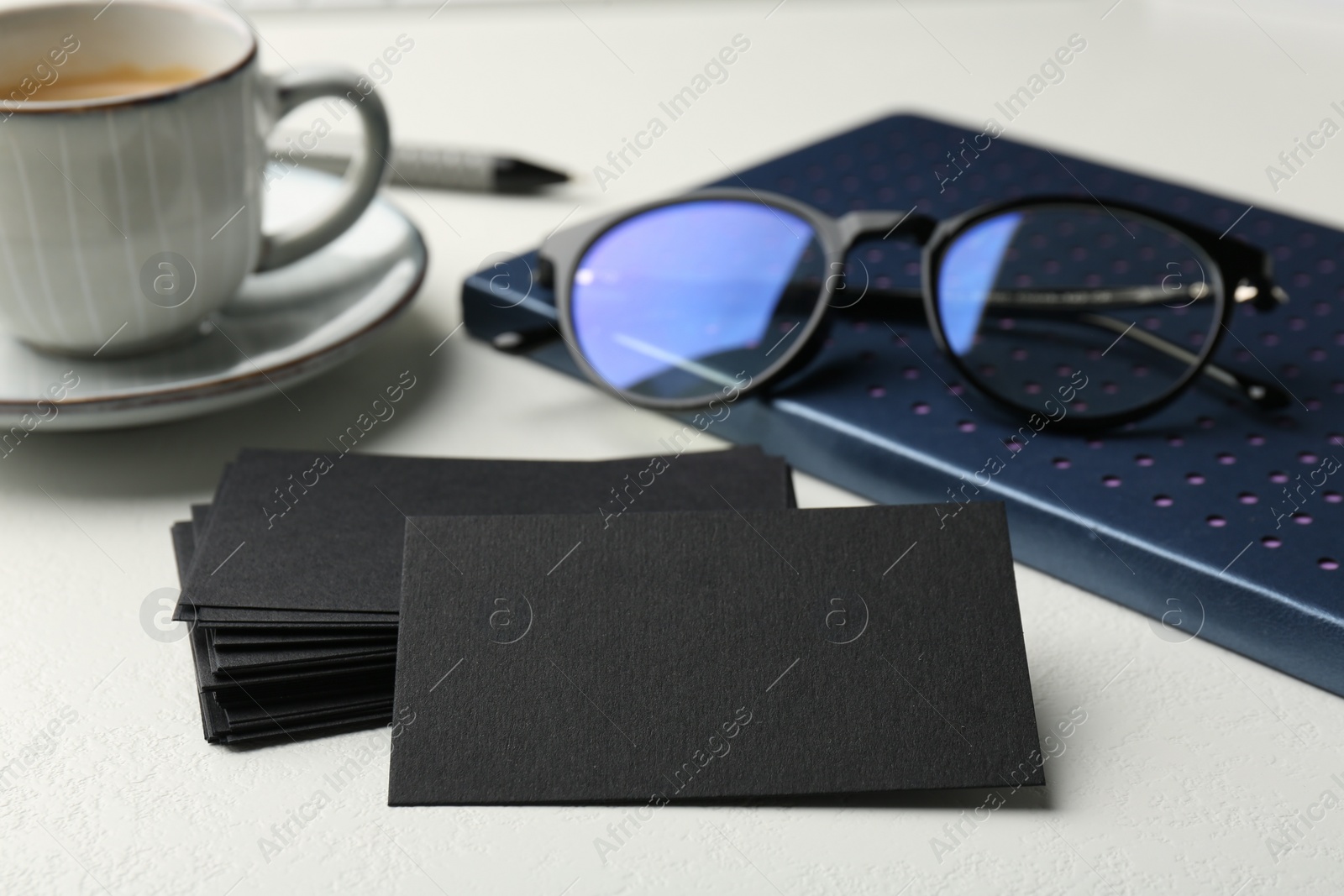 Photo of Blank black business cards, notebook, glasses and cup of coffee on white table, closeup. Mockup for design