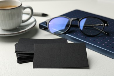 Photo of Blank black business cards, notebook, glasses and cup of coffee on white table, closeup. Mockup for design