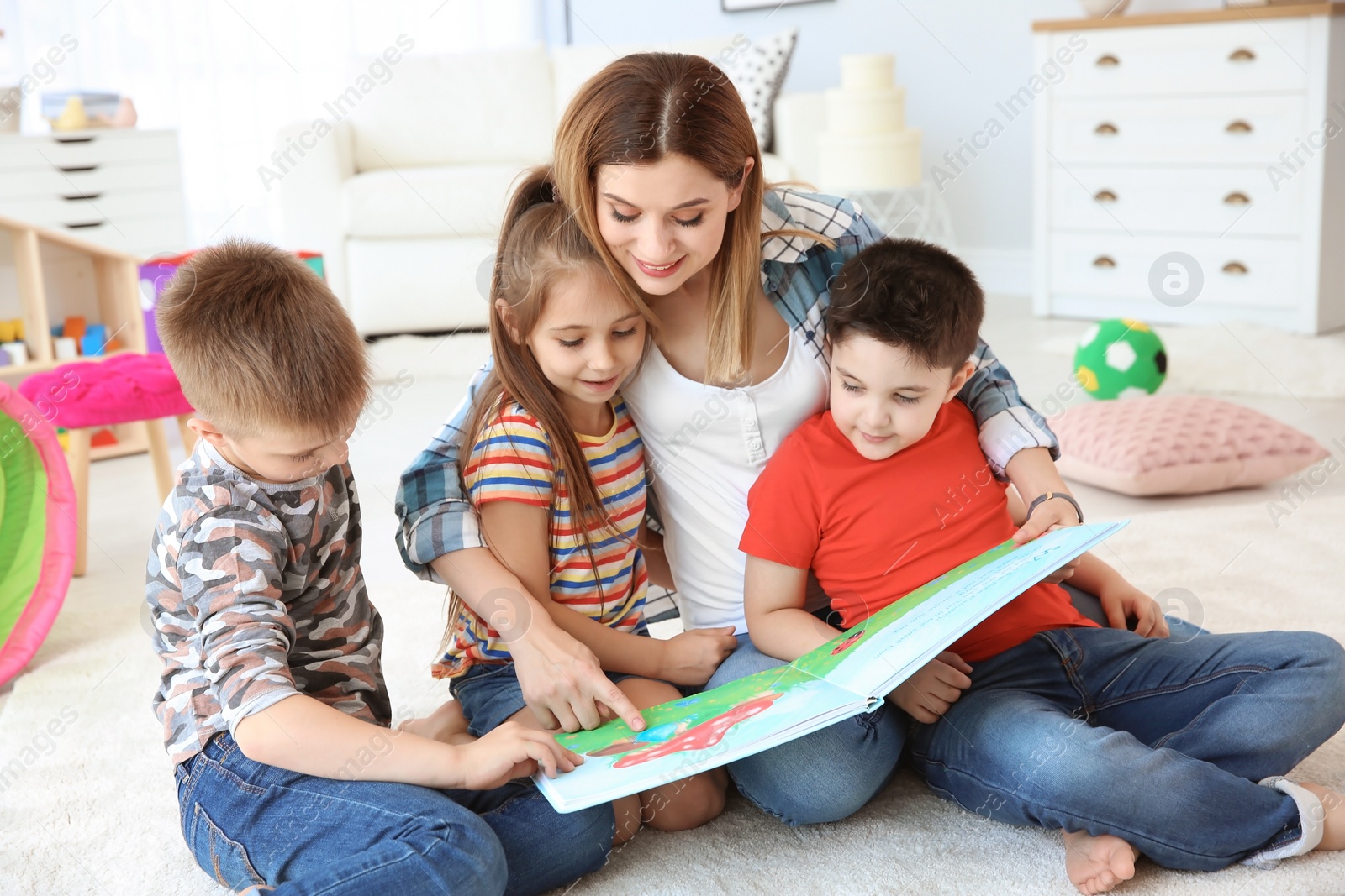 Photo of Cute little children reading book on floor with young mother in playing room