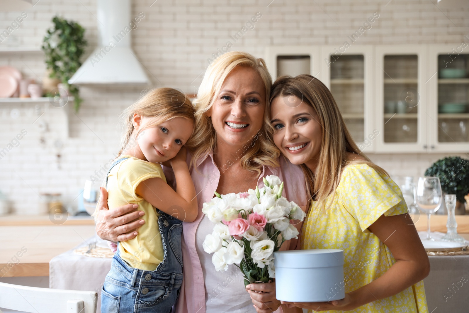 Photo of Young woman holding flowers and gift box with her mother and daughter in kitchen
