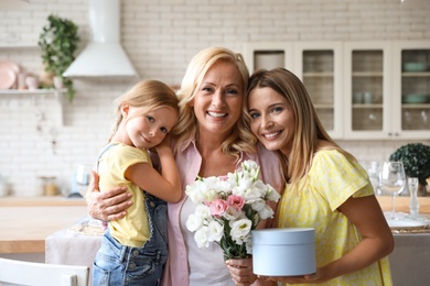 Photo of Young woman holding flowers and gift box with her mother and daughter in kitchen