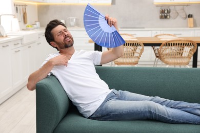 Bearded man waving blue hand fan to cool himself on sofa at home