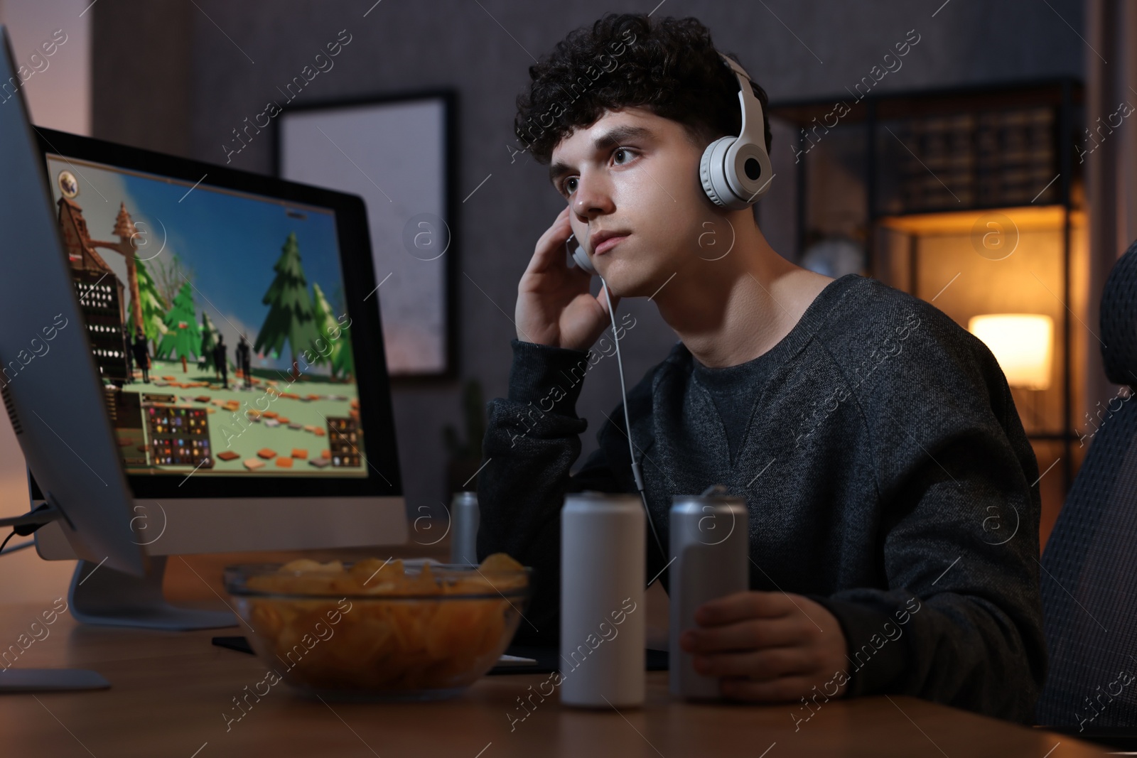 Photo of Young man with energy drink and headphones playing video game at wooden desk indoors