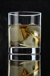 Photo of Tasty whiskey and ice cubes in glass on mirror table against black background, closeup