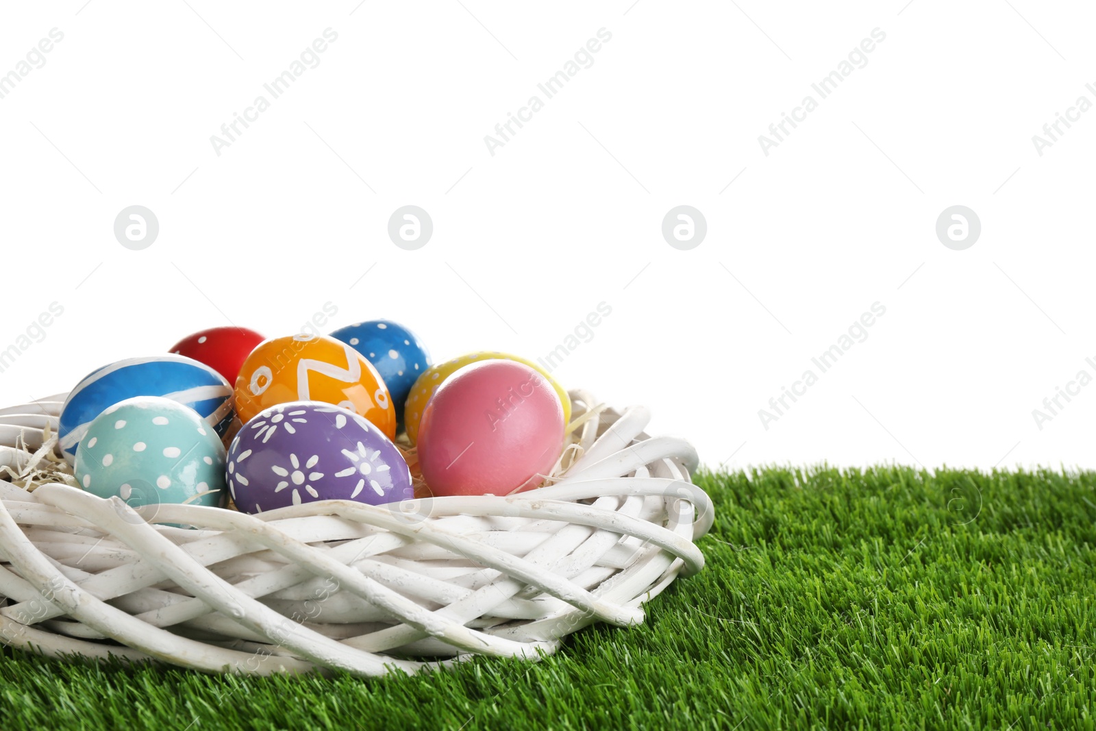 Photo of Wicker nest with painted Easter eggs on green grass against white background, space for text