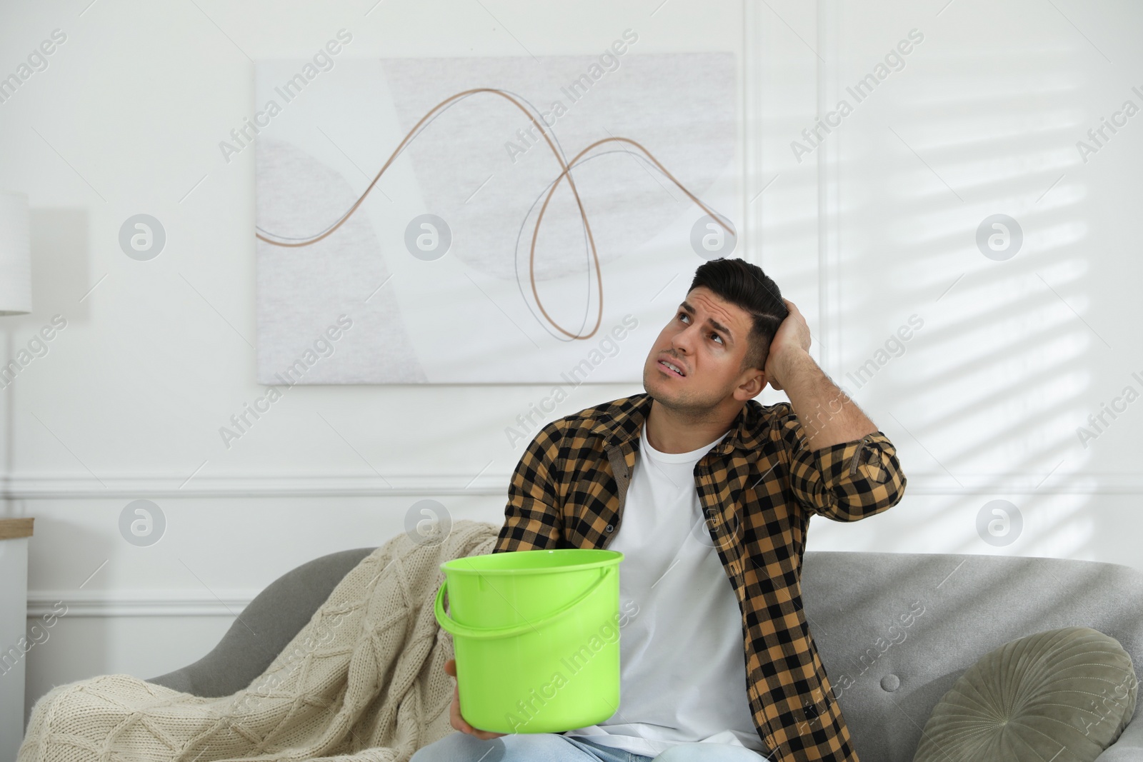 Photo of Emotional man collecting water leaking from ceiling in living room. Damaged roof