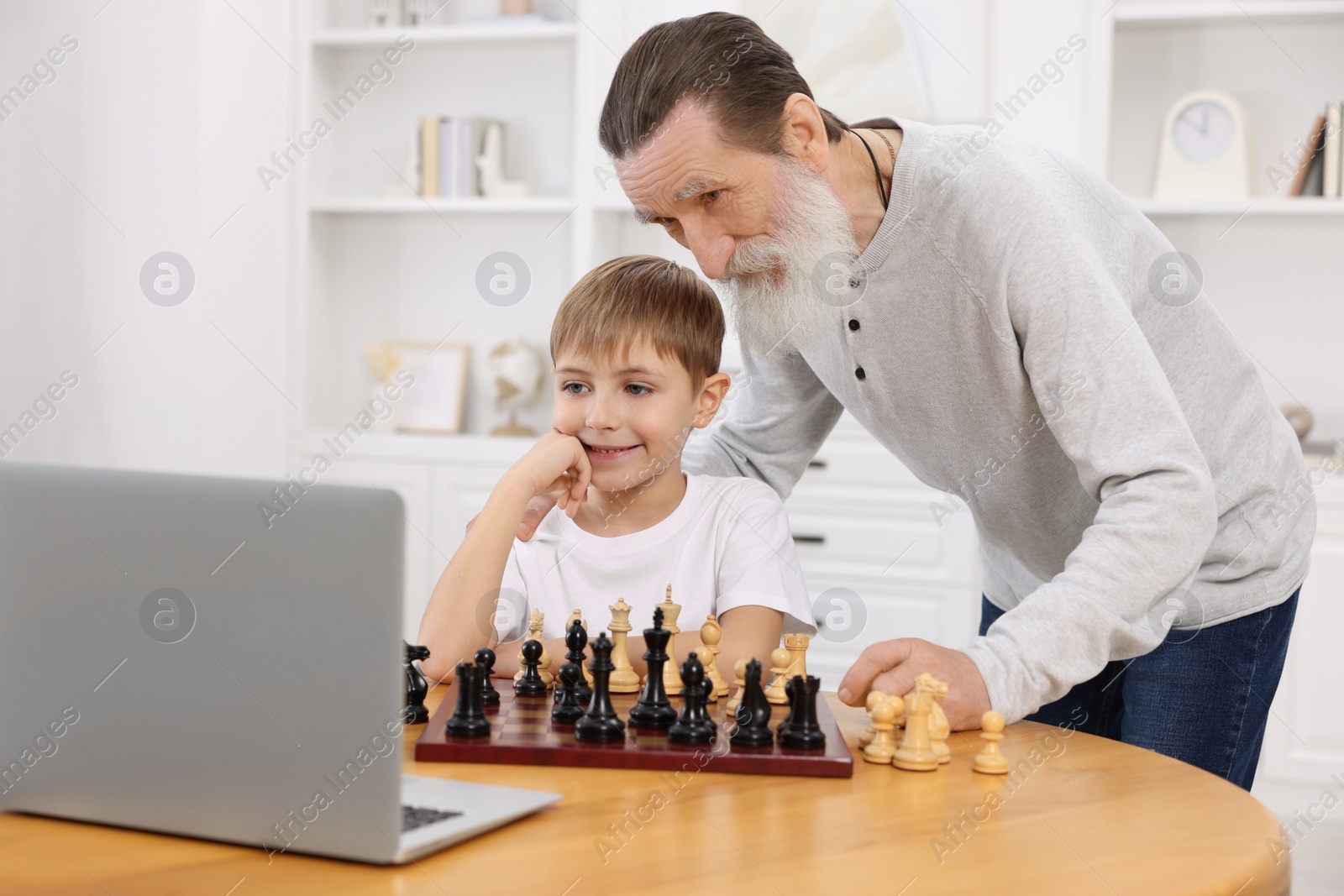 Photo of Grandfather teaching his grandson to play chess following online lesson at home