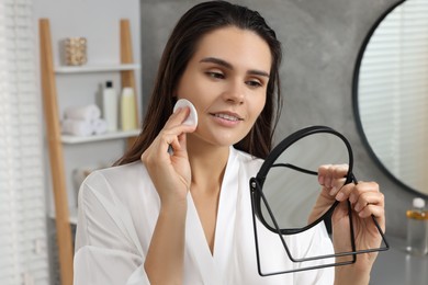 Photo of Young woman cleaning her face with cotton pad near mirror in bathroom