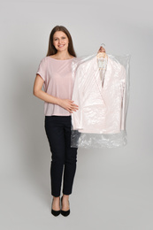 Young woman holding hanger with jacket in plastic bag on light grey background. Dry-cleaning service