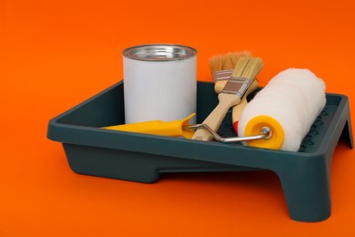 Can of orange paint, brushes, roller and container on color background