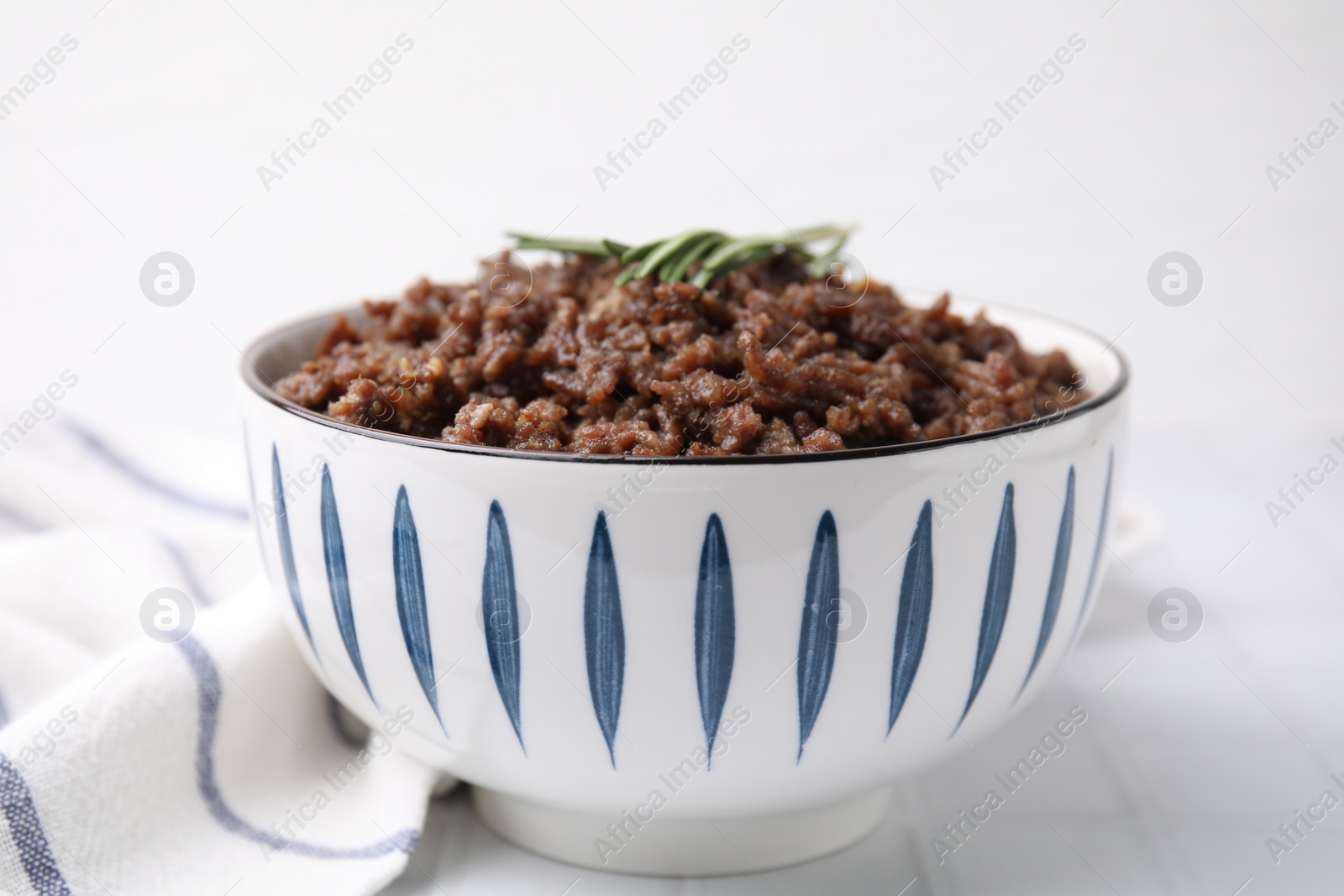 Photo of Fried ground meat in bowl and rosemary on white tiled table, closeup