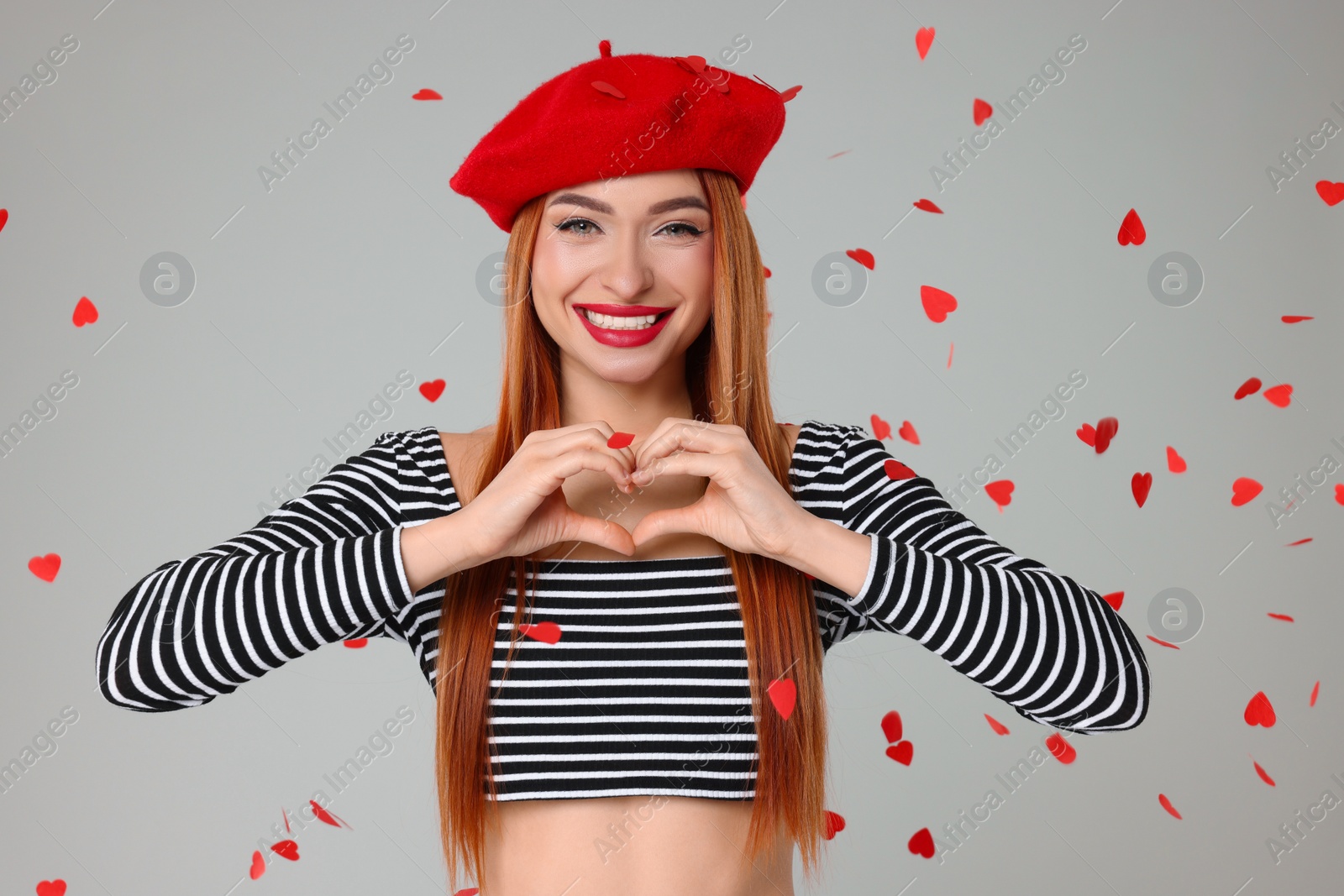 Photo of Young woman making heart with hands under confetti on light grey background